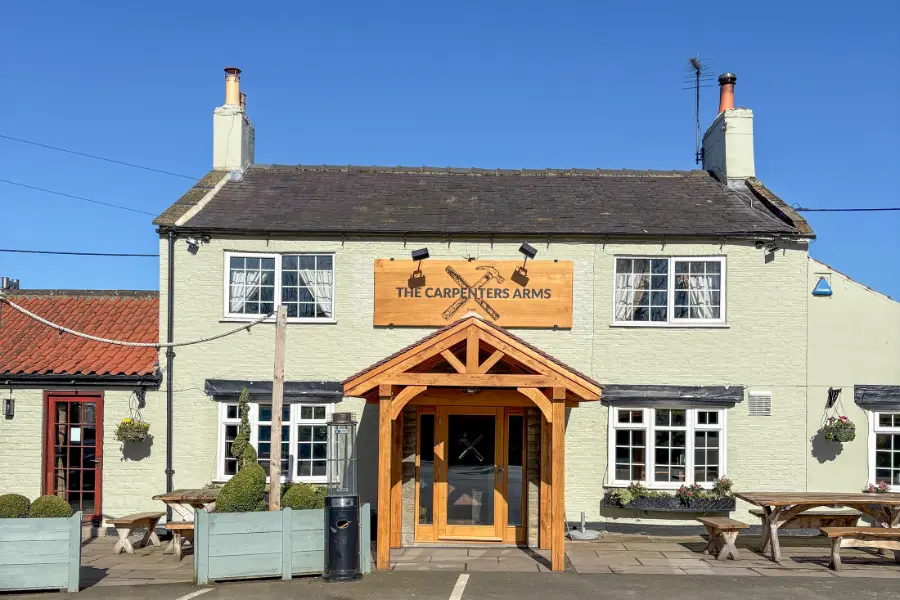 The front of the Carpenters Arms, Felixkirk, a cream painted village pub with a timber porch and picnic benches outside